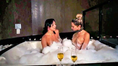 Two trans women in bubbly bath, nude with champagne glasses