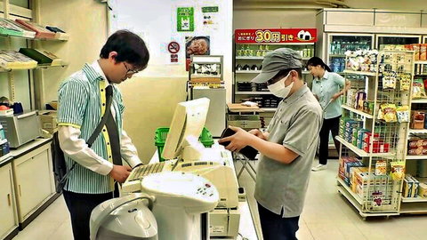 Two young Asian men wearing masks at convenience store checkout counter with snacks and drinks on shelves