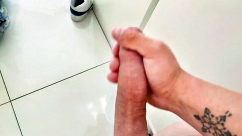 Tattooed hands gripping and stroking thick erect cock on tiled floor closeup