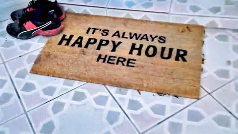 Doormat with 'It's Always Happy Hour Here' text and pink running shoes on tiled floor