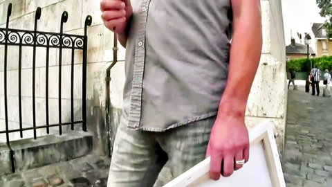 Muscular man in gray tank top and pants holding white frame against wrought iron fence on cobblestone street