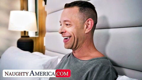 Smiling man in gray shirt grinning excitedly on luxury bedroom bed Naughty America