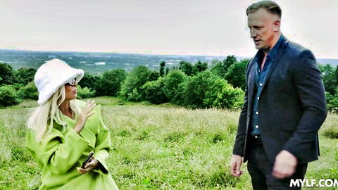Blonde MILF in green coat chats with Luke Hardy in outdoor field