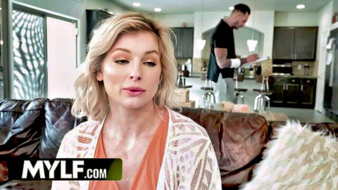 Blonde MILF in lacy orange top sits on couch with man in background