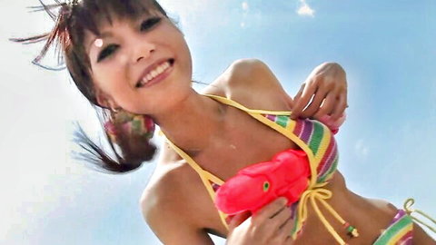 Skinny Japanese teen with small tits in colorful striped bikini holds red water gun to chest smiling outdoors