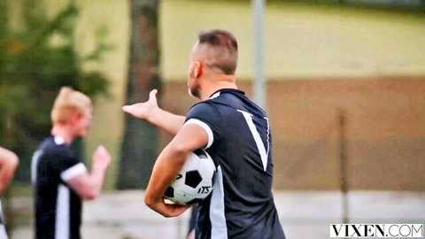 Muscular soccer player in tight black jersey holding soccer ball gesturing to blonde teammate on field