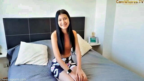 Petite Asian teen camgirl with small tits sitting topless on bed smiling seductively