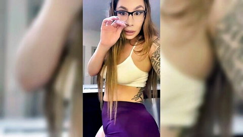 Tattooed Latina with long hair and glasses posing in white top and purple leggings