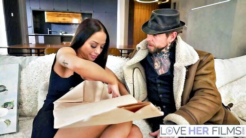 Brunette in black dress shares gift with tattooed bearded man in cozy scene