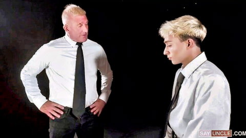 Blond daddy in suit stands dominantly facing submissive twink nephew in white shirt and tie