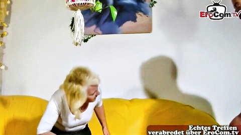 Mature blonde in white top and black pants leans seductively on yellow sofa teasing viewer