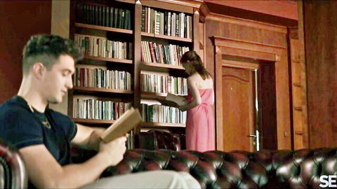 Handsome man reads book on leather chair as sexy brunette in red dress browses library shelves