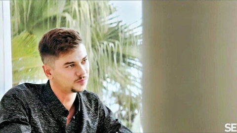 Handsome mustached man in open patterned shirt gazes sideways by palm tree window