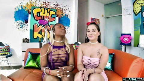 Ebony and brunette babes in sheer tops and chokers teasing tits on colorful couch