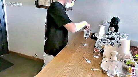 Man in black shirt and mask pouring white liquid from glass into another at wooden bar counter