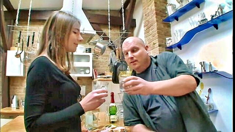Brunette woman in black dress toasts wine with bald master in BDSM kitchen scene
