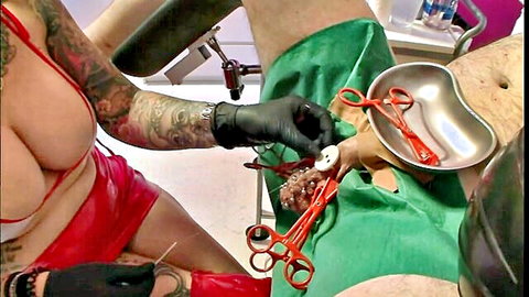 Tattooed femdom mistress in red latex spreads sub's exposed balls with speculum and red clamps in intense BDSM medical play