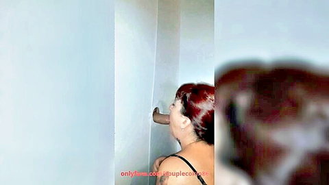 Redhead woman kneels at glory hole with stranger's hand reaching toward her mouth