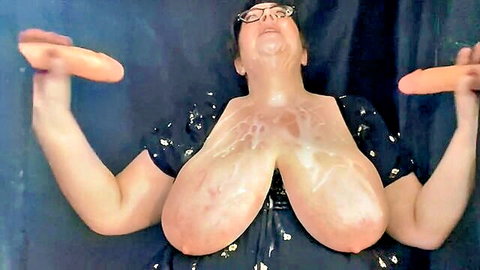 BBW with massive cum-covered tits holding two orange dildos in solo tease