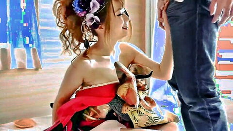 Smiling young Japanese babe in red draped kimono sits bare-shouldered holding mask as guy touches her