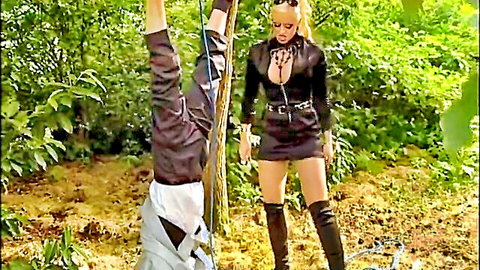 Blonde dominatrix in leather outfit binds upside-down man in forest BDSM