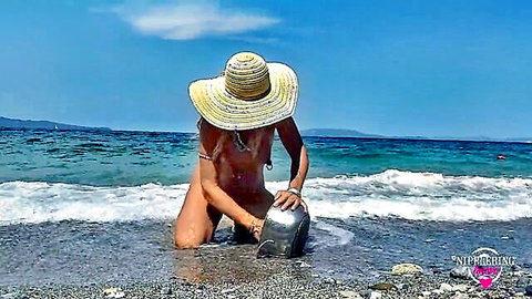 Skinny nude blonde with pierced nipples kneels on pebbly beach dipping silver helmet in waves exposing ass and pussy