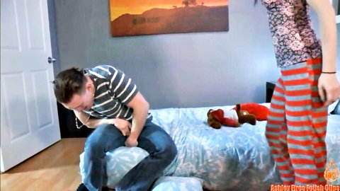 Ashley Fires in floral top and striped pants stands over guy gripping cock through jeans on bed