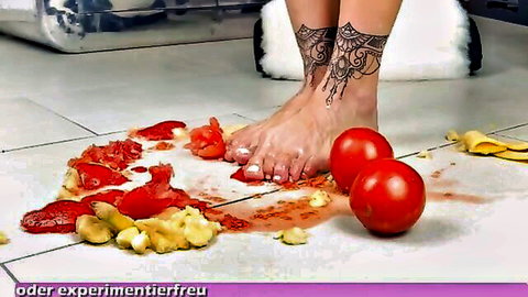 Tattooed bare feet with pink toes crush juicy tomatoes and bananas on floor in foot fetish video