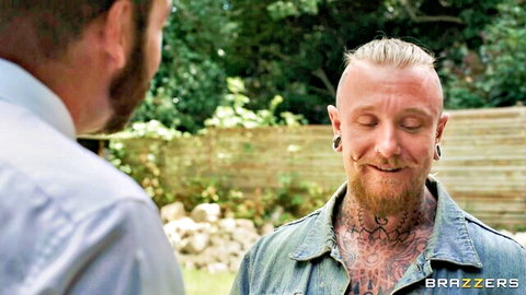 Tattooed blonde muscular guy with piercings smiling at suited bearded man outdoors in garden