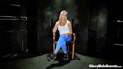 Blonde MILF with big tits in white tank top and blue leggings sits smiling in glory hole booth