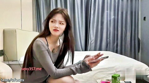 Asian beauty in gray top on hotel bed, teasing with phone