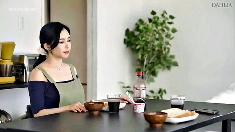 Beautiful Asian woman in off-shoulder top and green apron sitting at kitchen table with coffee and food
