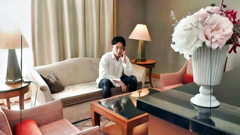 Handsome East Asian man in white shirt sits on hotel sofa checking phone amid flowers