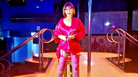 Curvy mature stripper in open pink satin robe revealing black lingerie and stockings on club stool by pole