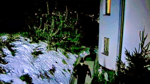 Mysterious hooded man in black coat walking up snowy steps at night toward lit white house