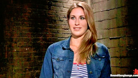 Blonde woman in denim jacket smiling against brick wall for gang anal DP preview