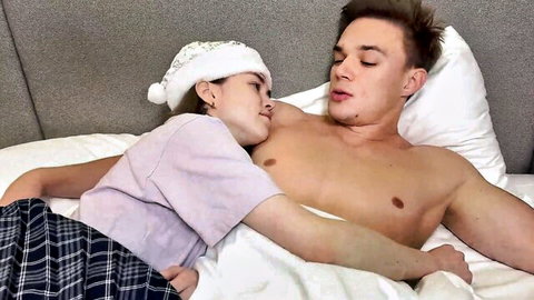 Petite Santa hat girl cuddles and kisses shirtless muscular guy's bare chest on bed