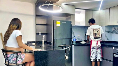 Sexy Latina in tiny shorts and crop top sits at kitchen counter watching guy cook