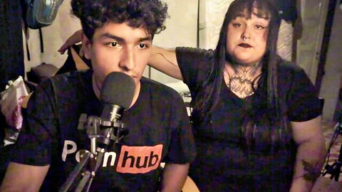 Chubby tattooed goth BBW stands behind curly-haired guy in Pornhub shirt speaking into mic