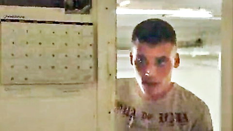 Young fair-skinned man with short hair stands by metal door in locker room wearing gray t-shirt
