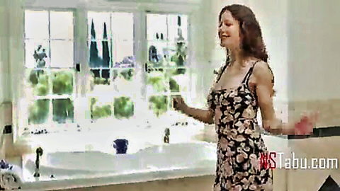 Curly-haired brunette stepmom in low-cut floral dress smiles seductively in bathroom