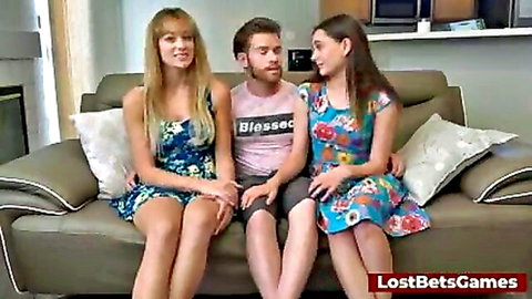 Smiling blonde and brunette in floral dresses with guy on couch, threesome tease