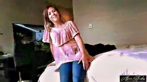 Smiling brunette wife in pink ruffled top and jeans on bed, teasing for cheating hotel affair