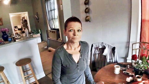 Short-haired stepmom in gray V-neck top, standing indoors for debt payment scene