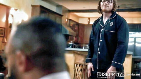 Young glasses-wearing brunette in black pajamas stands facing older man from behind in kitchen gay tease
