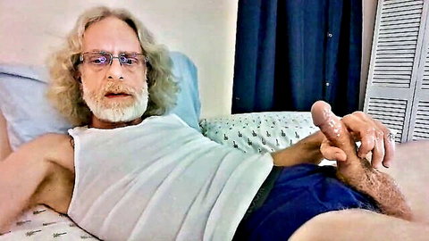 Skinny mature DILF with beard and glasses jerks his thick erect cock on bed in tank top
