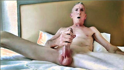 Naked old man spreads legs wide on bed gripping his thick erect cock and balls