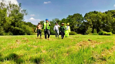 Group of muscular men in hi-vis vests and hard hats walking across grassy field outdoors