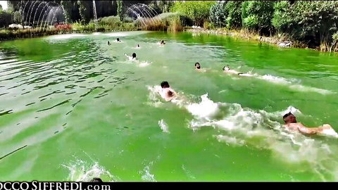 Group of swimmers splashing in vibrant green outdoor pond with fountains