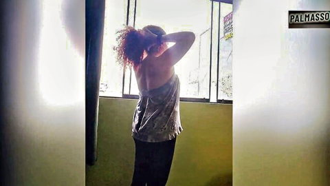 Mature curly redhead in sparkly top poses by window from behind